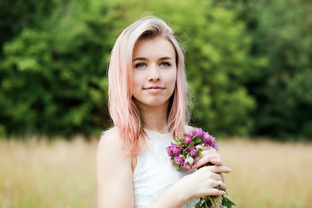 Schöne junge Frau in einem weißen Vintage-Spitzenkleid mit rosa Haaren und einem Hut geht über die Wiese