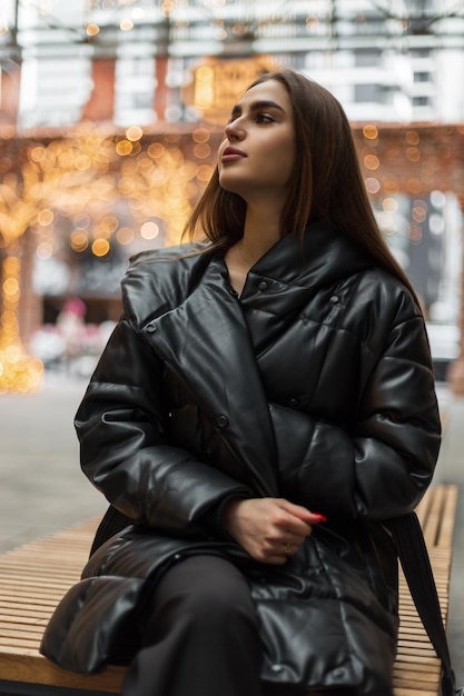 Schöne junge Frau in einer modischen Winterjacke sitzt auf einer Bank in der Stadt in der Nähe der gelben Lichter