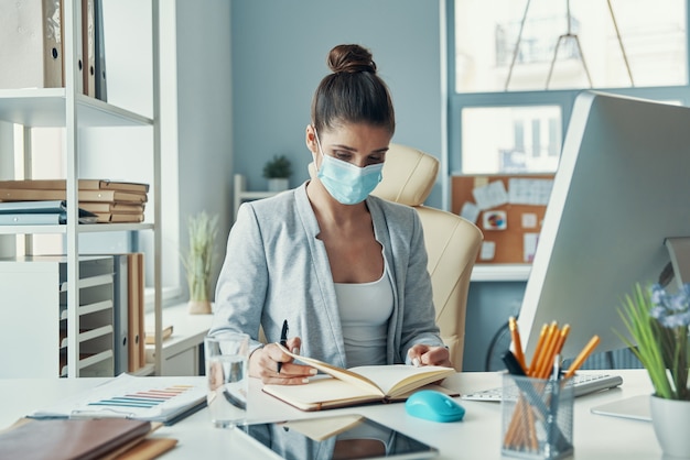 Schöne junge Frau in eleganter Freizeitkleidung, die etwas aufschreibt, während sie im Büro sitzt