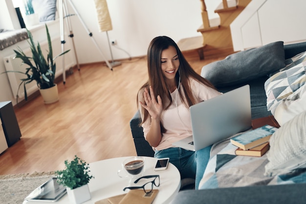 Schöne junge Frau in Freizeitkleidung, die mit jemandem spricht, der einen Laptop benutzt, während sie sich zu Hause ausruht