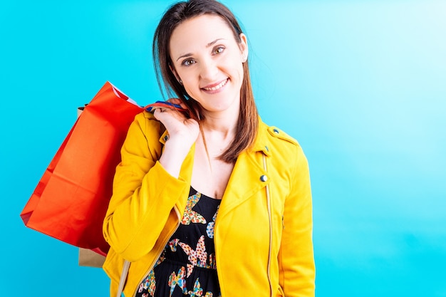 Schöne junge Frau in gelber Jacke auf blauem Hintergrund mit bunten Einkaufstaschen. sie shopaholic Konzept. Shopping Wochenende. Verkauf