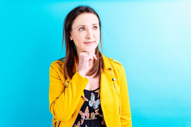 Schöne junge Frau in gelber Jacke auf blauem Hintergrund nachdenklich mit Exemplar beiseite schauend