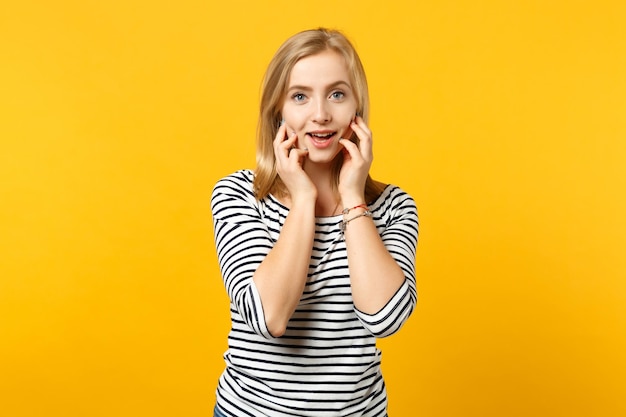 Schöne junge Frau in gestreifter Kleidung, die in die Kamera schaut und die Hände auf die Wangen legt, isoliert auf gelb-orangefarbenem Wandhintergrund im Studio. Menschen aufrichtige Emotionen, Lifestyle-Konzept. Mock-up-Kopienbereich.