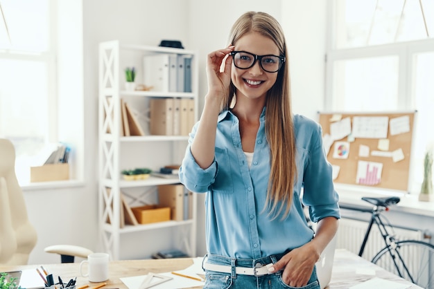 Schöne junge Frau in intelligenter Freizeitkleidung, die Kamera anschaut und lächelt, während sie im Büro steht