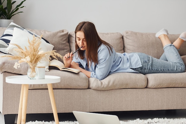 Schöne junge Frau in Jeans und blauem Hemd, die sich mit Notizblock und Stift am Sofa hinlegt