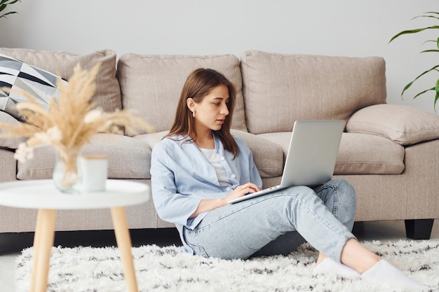 Schöne junge Frau in Jeans und blauem Hemd sitzt zu Hause mit Laptop auf dem Boden