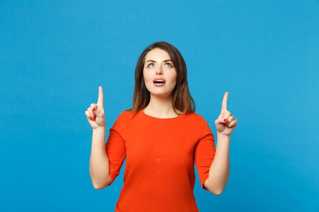 Schöne junge Frau in rot-orangefarbenem Kleid, die posiert und mit den Fingern auf den Arbeitsplatz zeigt, isoliert über trendigem blauem Wandhintergrund, Studioporträt. People-Lifestyle-Konzept. Mock-up-Kopienbereich.