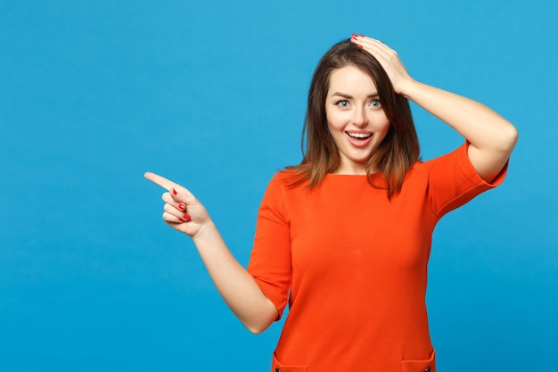 Schöne junge Frau in rot-orangefarbenem Kleid, die posiert und mit den Fingern auf den Arbeitsplatz zeigt, isoliert über trendigem blauem Wandhintergrund, Studioporträt. People-Lifestyle-Konzept. Mock-up-Kopienbereich.