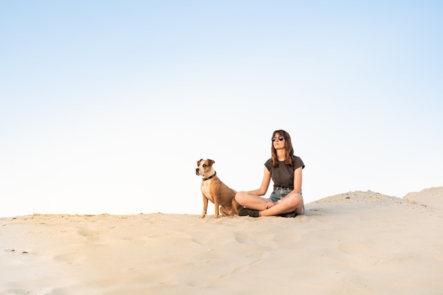 Schöne junge Frau in Sonnenbrille mit Hund sitzen auf Sand. Mädchen im Wandern Freizeitkleidung und Staffordshire Terrier Welpe sitzen am Sandstrand oder in der Wüste an heißen sonnigen Tag