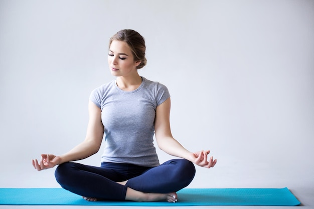 Schöne junge Frau in Sportkleidung, die Yoga praktiziert, während sie im Lotussitz auf grauem Hintergrund sitzt Yoga-Fitness und Training