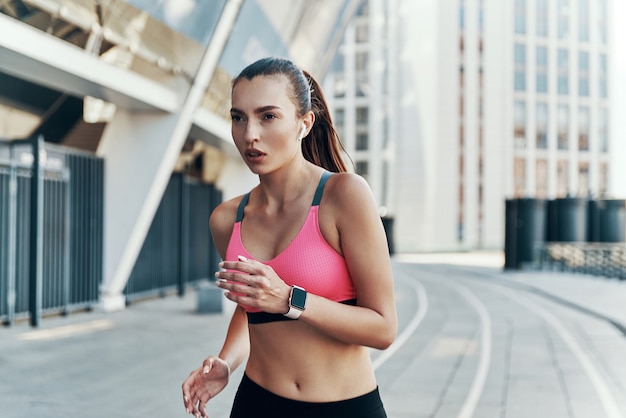 Schöne junge Frau in Sportkleidung mit Kopfhörern und Smartwatch beim Laufen im Freien