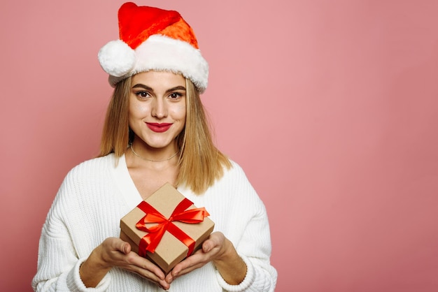 Schöne junge Frau in Weihnachtsmütze hält ein Weihnachtsgeschenk lächelnd auf einem rosa Hintergrund