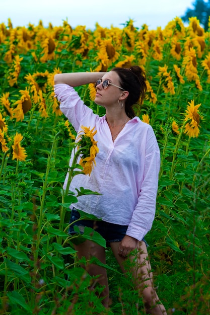 Schöne junge Frau in weißem Hemd und Sonnenbrille, die an einem Sommertag zwischen gelben Sonnenblumen posiert.