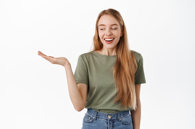 Schöne junge Frau lächelt, schaut ihre leere offene Hand erfreut an, trägt einen Artikel, zeigt Werbeprodukt in der Handfläche an und steht im T-Shirt gegen weiße Wand