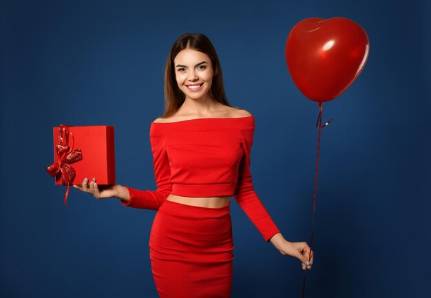 Schöne junge Frau mit Ballon und Geschenk zum Valentinstag auf farbigem Hintergrund