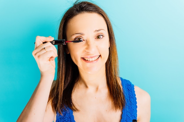 Schöne junge Frau mit blauem Hintergrund, die sich Wimperntusche auf ihren Wimpern lächelt