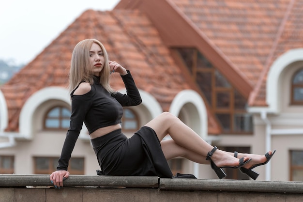 Schöne junge Frau mit blonden Haaren trägt schwarze Kleidung, langen Rock und High Heels. Stilvolles Mädchen draußen. Altes Gebäude im Hintergrund
