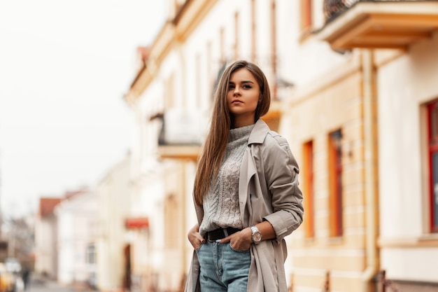 Schöne junge Frau mit braunen langen Haaren in einem stilvollen Trenchcoat, der in der Stadt draußen aufwirft