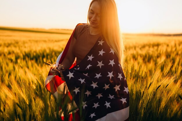 Schöne junge Frau mit der amerikanischen Flagge in einem Weizenfeld bei Sonnenuntergang