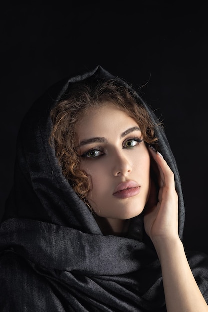 Schöne junge Frau mit dunkelgrauem Kopftuch bedeckt ihr Gesicht mit luxuriösem Make-up auf schwarzem Hintergrund, der nach unten schaut