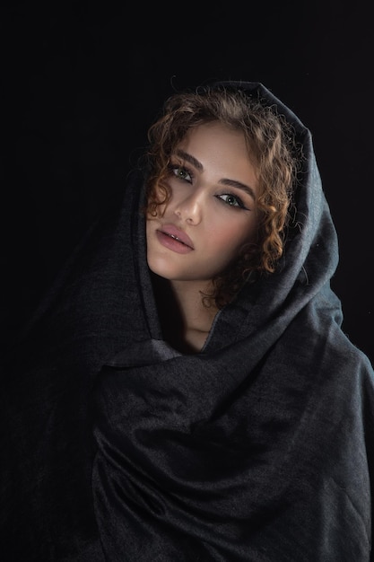 Schöne junge Frau mit dunkelgrauem Kopftuch bedeckt ihr Gesicht mit luxuriösem Make-up auf schwarzem Hintergrund, der nach unten schaut