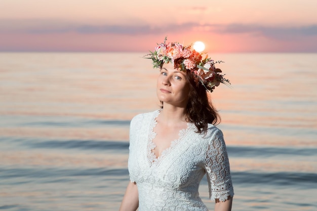 Schöne junge Frau mit einem Blumenkranz auf dem Kopf am Meer bei Sonnenuntergang