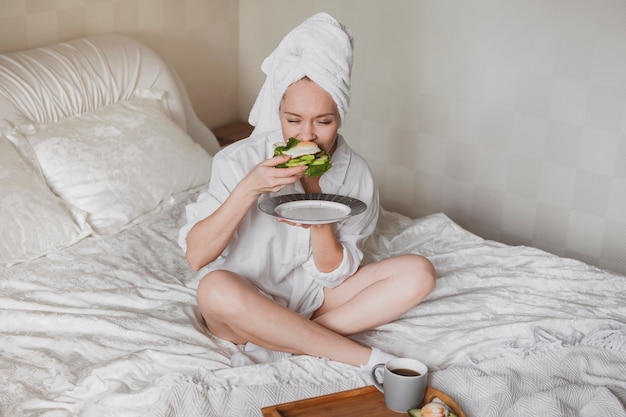 Schöne junge Frau mit einem Handtuch auf dem Kopf im Bett, das gesundes Frühstück isst