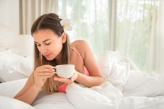 Schöne junge Frau mit einem heißen Getränk in einem hellen Bett. Entspannung und fauler Morgen Konzept.