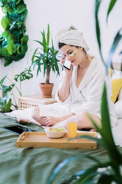 Foto schöne junge frau mit goldenen augenklappen, mobiltelefon sitzt auf dem bett im bademantel und handtuch im schlafzimmer zu hause, selbstpflege, anti-age-behandlung für die haut, surfen, online-shopping, coronavirus-isolation