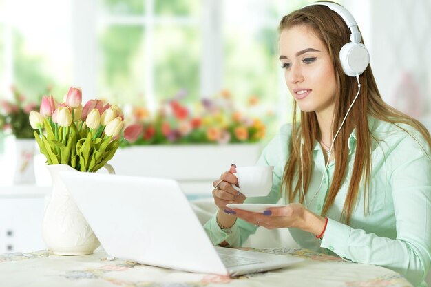 Schöne junge Frau mit Kopfhörern, die am Tisch sitzt und Laptop verwendet