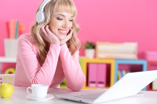 Schöne junge Frau mit Kopfhörern mit Laptop im Zimmer mit rosa Wänden