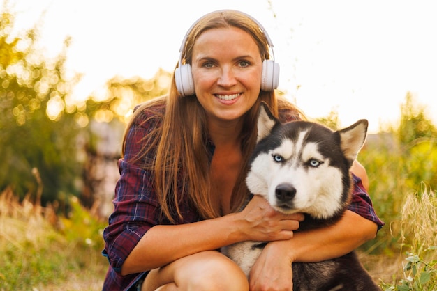 Schöne junge Frau mit Kopfhörern spielt bei Sonnenuntergang mit ihrem Hund im Freien Umarmungen