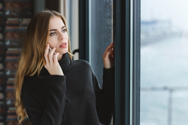 Schöne junge Frau mit langen Haaren, die aus dem Fenster schaut und am Handy spricht