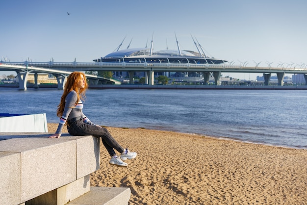 Schöne junge Frau mit langen Haaren kaukasischer Nationalität, in Freizeitkleidung sitzt an einem sonnigen Frühlingstag am Ufer des Flusses mit Blick auf die Brücke the