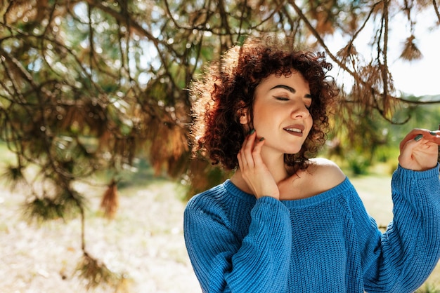 Schöne junge Frau mit lockigem Haar, die lächelt und das Wetter genießt, das vor Naturhintergrund posiert, hat einen positiven Ausdruck