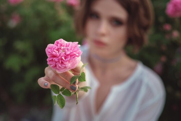 Schöne junge Frau mit lockigem Haar posiert in der Nähe von Rosen in einem Garten Das Konzept der Parfümwerbung