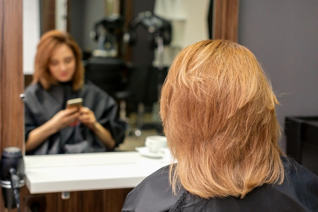 Schöne junge Frau mit roten Haaren, die ihr Smartphone benutzt und vor einem Spiegel sitzt und darauf wartet, einen Friseur zu besuchen.