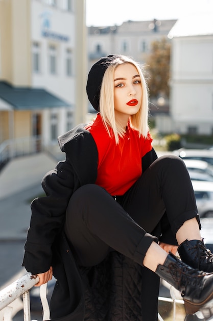 Schöne junge Frau mit roten Lippen in einem trendigen schwarzen Herbstmantel mit einer Baskenmütze auf der Straße an einem sonnigen Tag