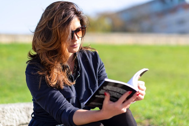 Schöne junge Frau mit schöner Frisur und Sonnenbrille, die an einem sonnigen Tag im Freien sitzt und ein Buch liest