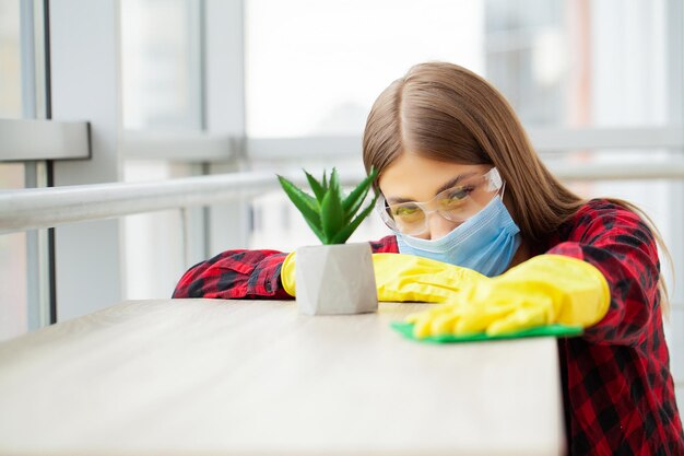 Schöne junge Frau mit Schutzhandschuhen, die den Schreibtisch im Büro putzt