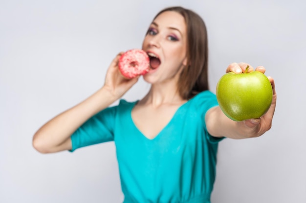 Schöne junge Frau mit Sommersprossen im grünen Kleid, die rosa Donut isst und grünen Apfel hält