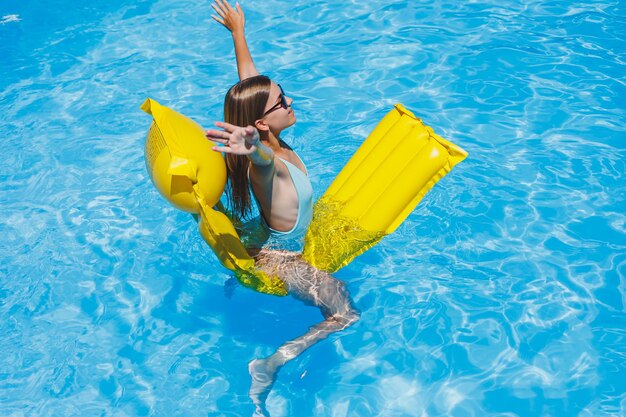 Schöne junge frau mit sonnenbrille auf einer gelben aufblasbaren matratze in einem badeanzug schwimmt im pool eines sommerferienreiselächelns eines luxushotels