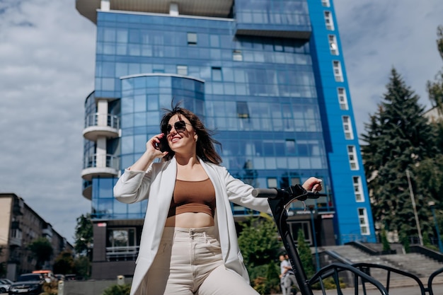 Schöne junge Frau mit Sonnenbrille und weißem Anzug, die auf ihrem Elektroroller in der Nähe eines modernen Gebäudes und Telefons steht