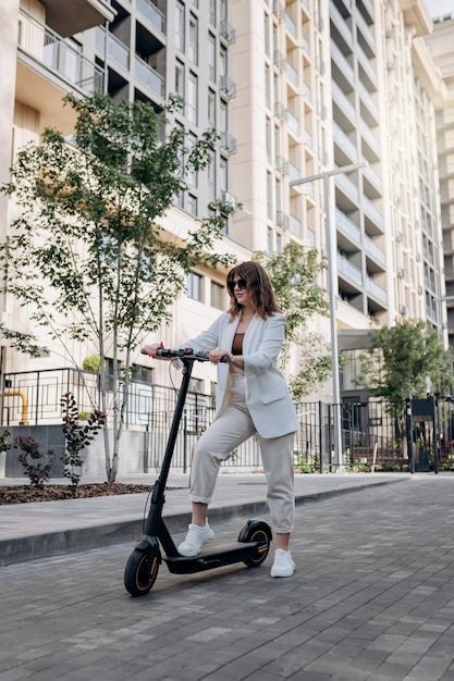 Schöne junge Frau mit Sonnenbrille und weißem Anzug, die mit ihrem Elektroroller in der Nähe eines modernen Gebäudes in der Stadt steht und wegschaut