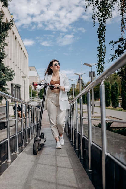 Schöne junge Frau mit Sonnenbrille und weißem Anzug, die mit ihrem Elektroroller in der Nähe eines modernen Gebäudes spazieren geht und wegschaut