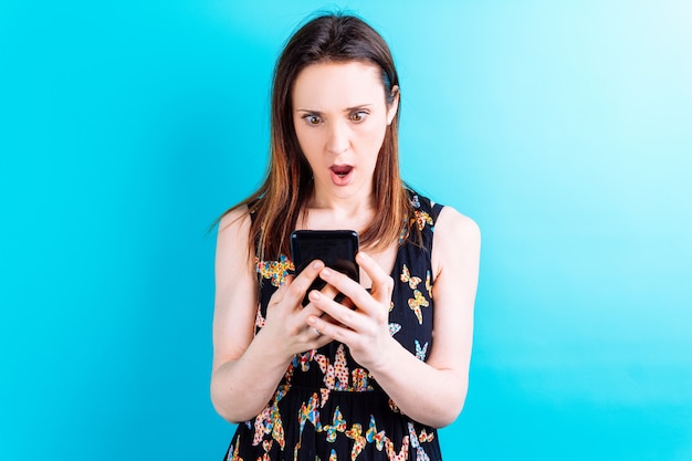 Schöne junge Frau mit überraschtem Gesicht, das Smartphone im blauen Hintergrund mit Exemplar betrachtet