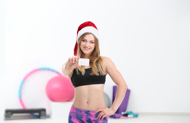 Schöne junge Frau mit Weihnachtsmann-Hut mit Karte im Fitnessstudio