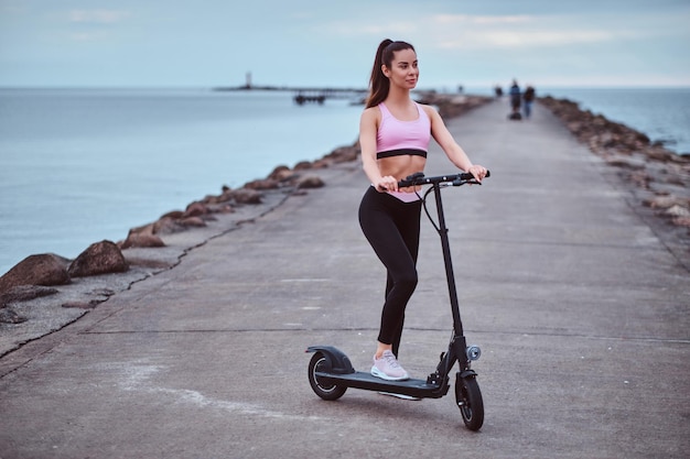 Schöne junge Frau posiert mit ihrem Elektroroller für Fotografen am Meer.