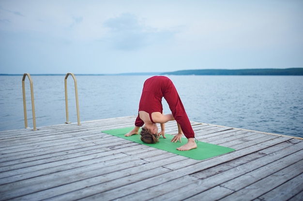 Schöne junge Frau praktiziert Yoga Asana Prasarita Padottanasana auf dem Holzdeck in der Nähe des Sees