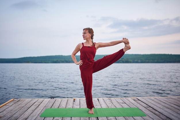 Schöne junge Frau praktiziert Yoga Asana Utthita Hasta Padangushthasana auf dem Holzdeck in der Nähe des Sees